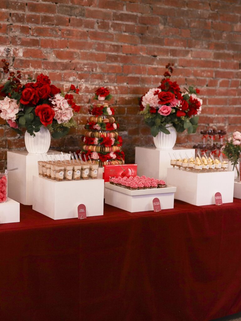 Gluten-free dessert installation by Kinn Studios for Ma Chérie bridal shower in Calgary, featuring pastel-pink and deep-red macaron tower, mini cheesecake shooters, mousse verrines, cherry-in-coupe glasses by Deep's Delights Catering in Calgary, and custom Café Rumie signage on a burgundy linen table against exposed brick