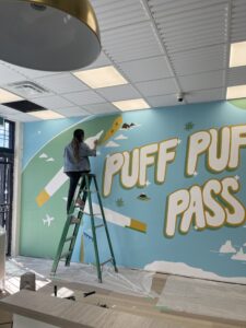 A colorful mural titled "Puff Puff Pass" by Calgary Mural Artist Kiran Rai-Bhullar, founder of Kinn Studios for Bud Mart Cannabis. This mural is a play on words that celebrates global cannabis culture.